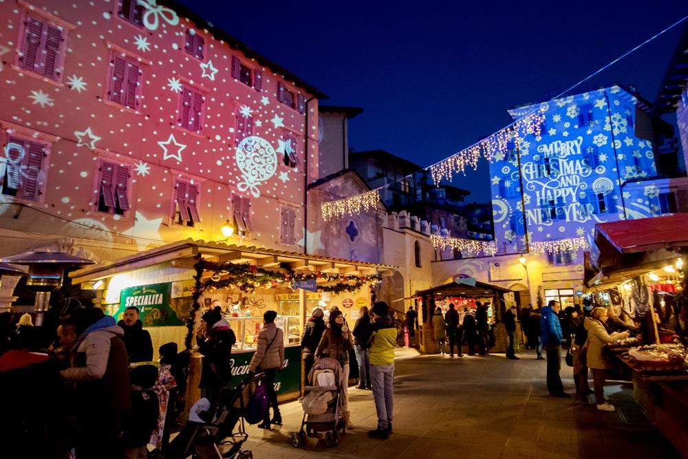 A holiday market in a city square with booths, fairy lights, and buildings lit up with Christmas projections.